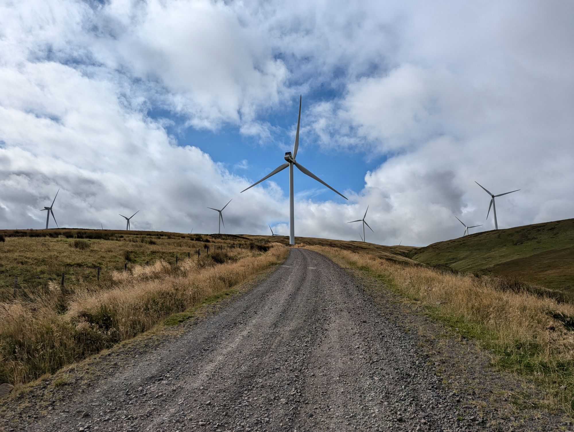 Gravel access track leading up to a wind turbine on Scottish hills, further turbines on the skyline under a bright broken sky